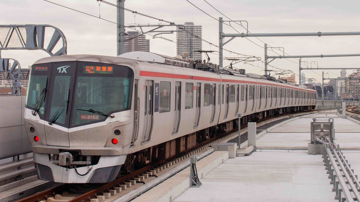 A sleek modern train traverses an elevated railway in an urban Japanese setting.